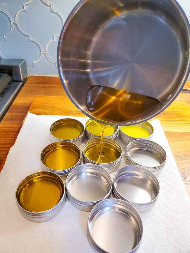 A stainless steel pot is poised over metal tins, pouring melted golden liquid into the tins below. 