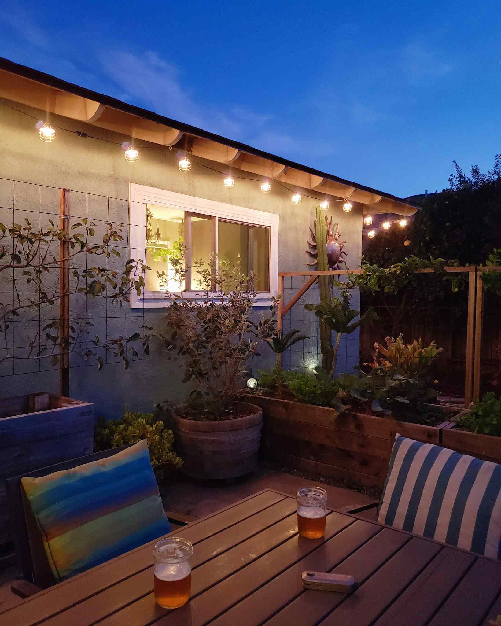 The backyard patio is shown during dusk. There are two beers on the patio table and the string lights that line the eaves of the house are lit. When one starts a homestead, it is a good idea to take time for yourself and relax on occasion. 