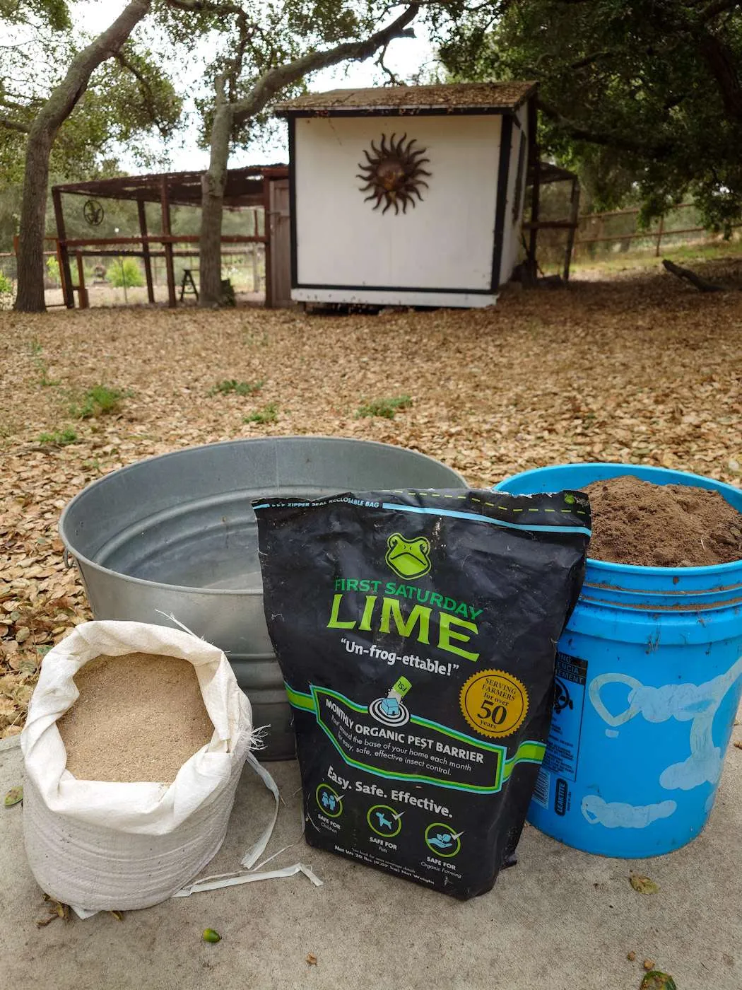A sand bag full of sand, a bag of lime, and a blue bucket of native soil sit next to each other in front of an empty metal tub. These are the makings of a chicken dust bath. Beyond there is a large coop and run where the chickens reside. 
