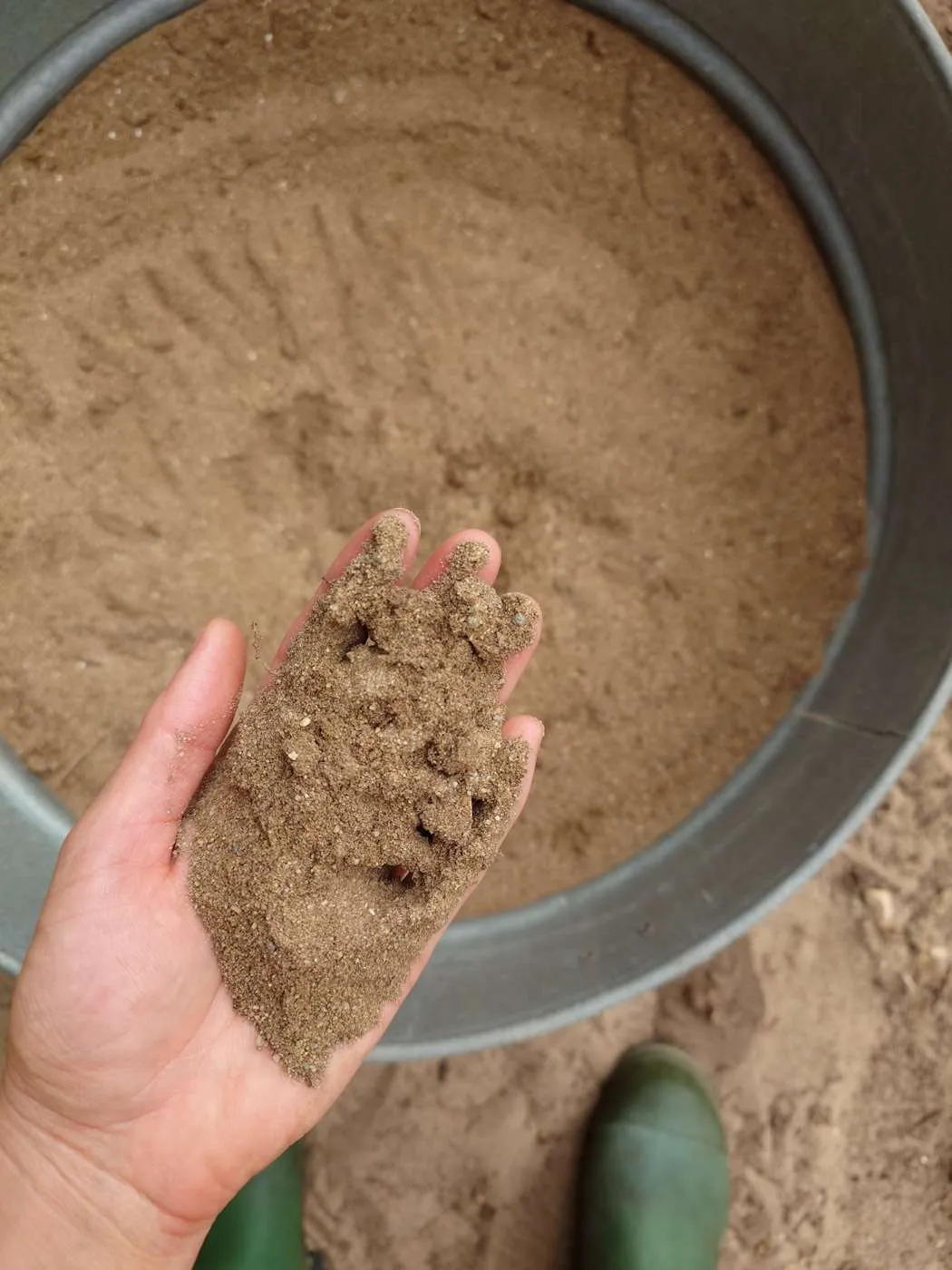 A hand is holding a handful of dirt and sand to illustrate the texture and makeup of the contents in the metal tub that is visible below. 