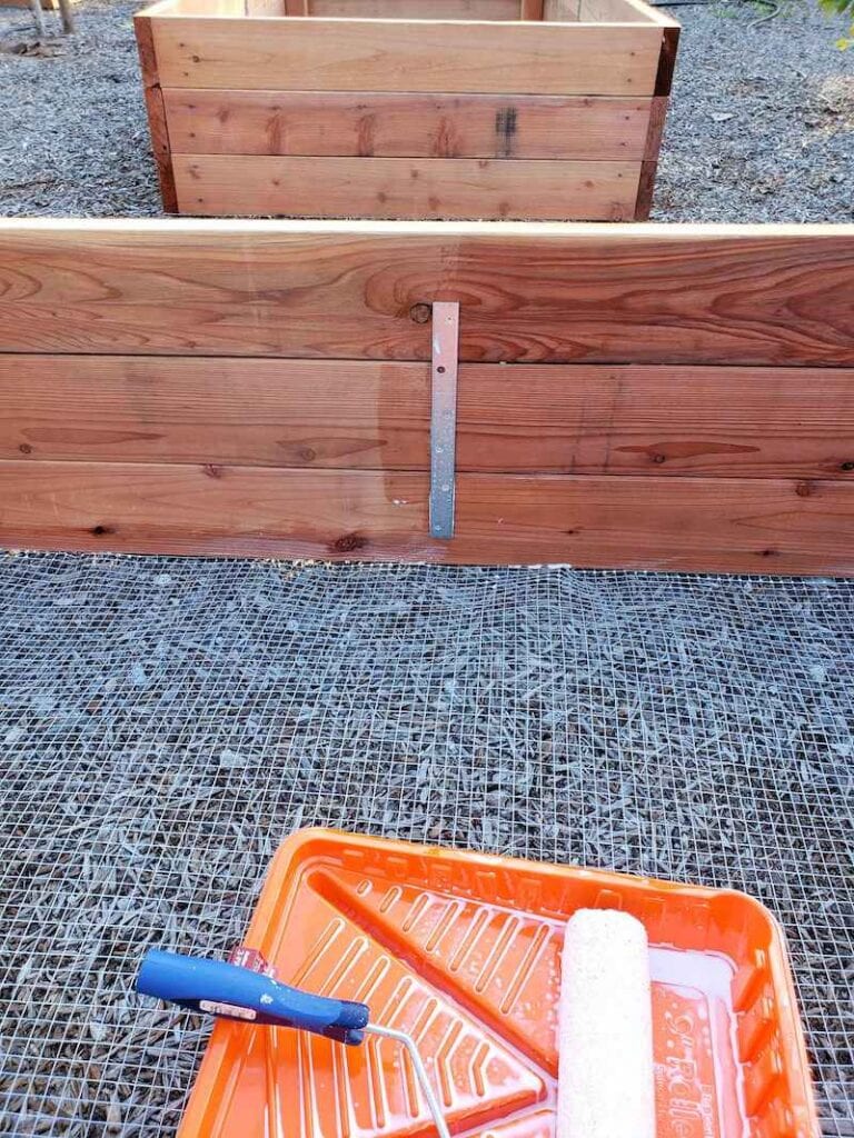 The inside of a garden bed is shown, half of the bed has had an application of sealant where the other side is still natural. The sealed side is slightly darker than the unsealed side. A paint tray and roller sit in the foreground with some milky colored sealant in the bottom. Seal beds to make garden beds last longer. 