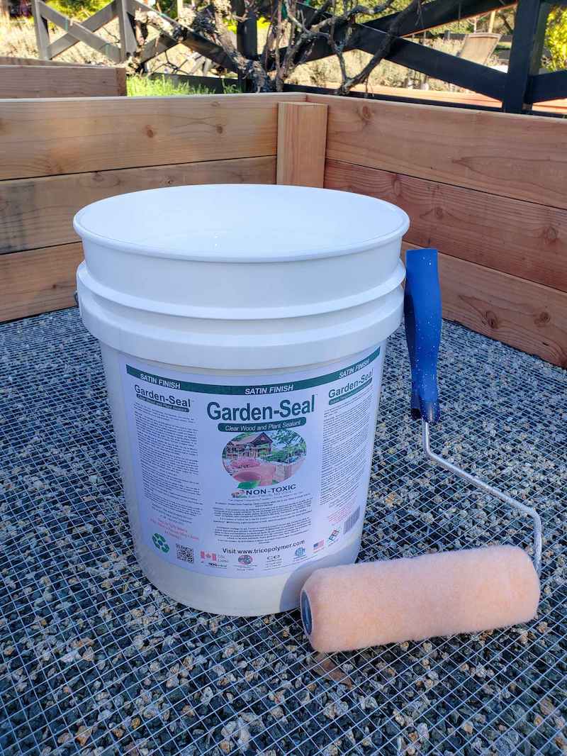 A 5 gallon bucket of Garden-Seal sits inside a raised bed. A paint roller is upside down, leaning against the bucket. 