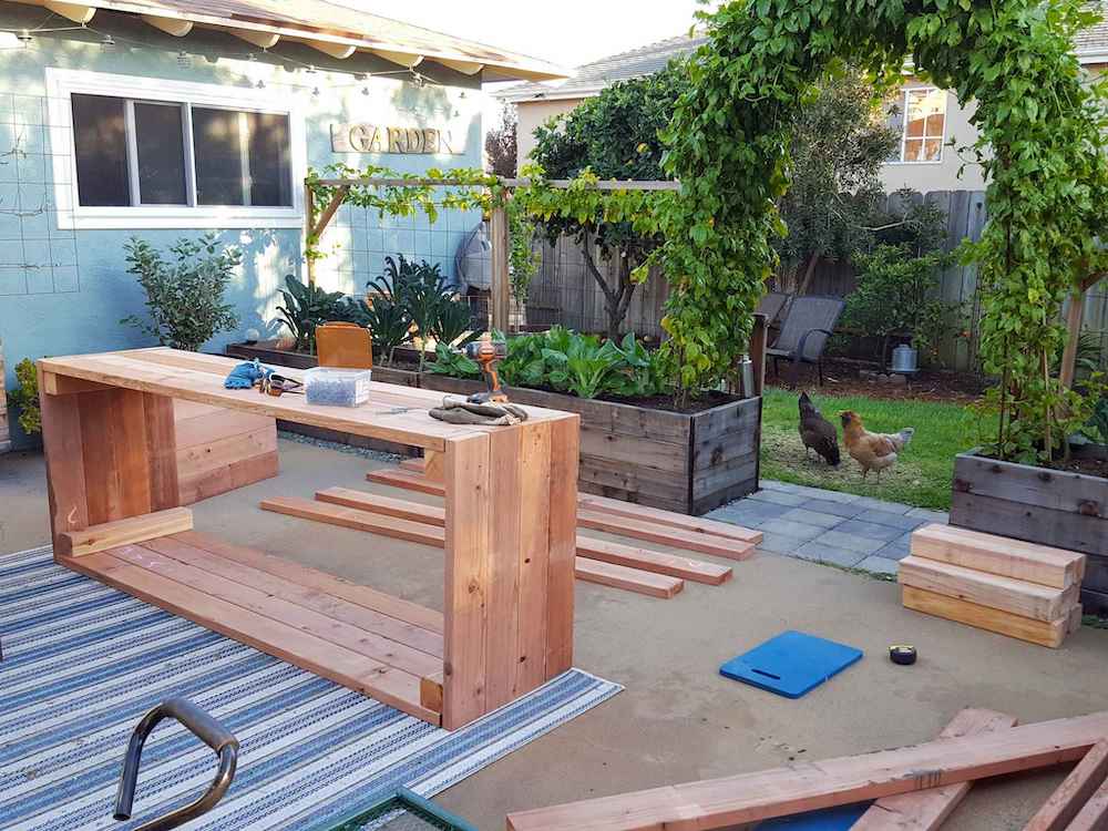 A concrete patio is being used to construct garden beds. There are pieces of 2x6 boards and 4x4 boards as well. One bed is laying on its side, fully constructed. The patio is lined with older garden beds that are filled with various vegetables. Two chickens stand in the yard beyond, visible between two of the beds that create a gate. Using cedar or redwood help make garden beds last longer. 