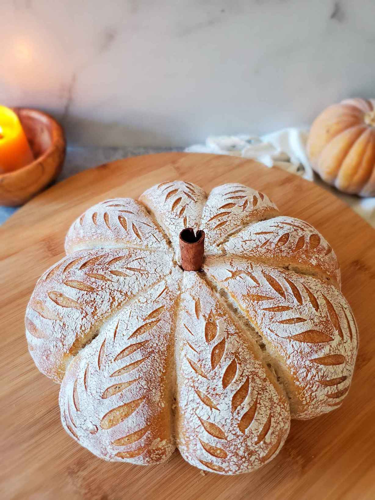 A baked loaf of pumpkin shaped sourdough bread sits atop a circular wooden board. A cinnamon stick has been inserted into the middle of the loaf to act as the stem. A small pumpkin and a candle sit just beyond the board as decor. 