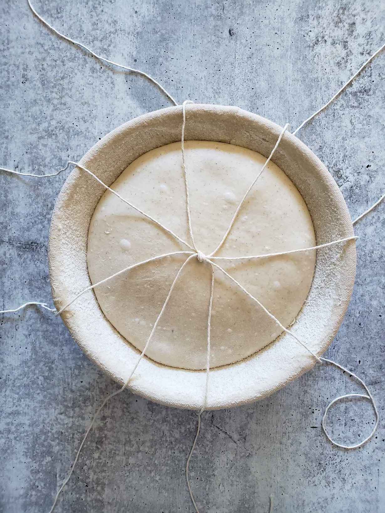 A loaf of dough is in a boule shaped banneton, pieces of string are splayed out into 8 sections like an octopus, all of them are connected in the middle by a knot. 