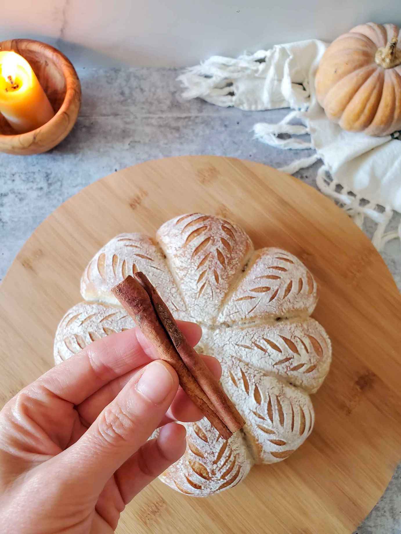 A hand is holding a cinnamon stick that will be used as a stem for the loaf of bread that sits below on a circular wood surface. 