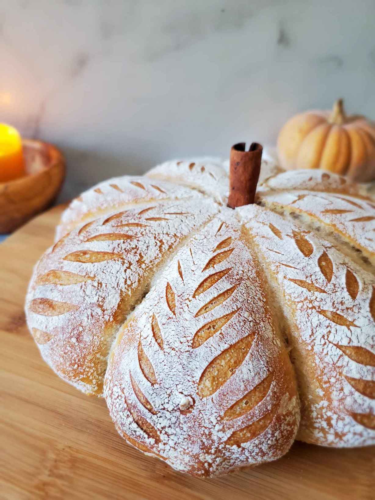 A close up image of half of a pumpkin shaped sourdough bread loaf. A cinnamon stick is inserted into the top to look like the stem. In the background there is a smaller real pumpkin and a small candle in a wooden bowl. 