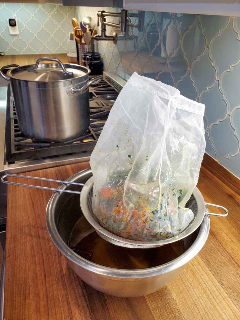 A fine mesh strainer is perched over a large metal bowl. Inside the strainer is a nut milk bag which contains dried chickweed and calendula flowers that have been strained out of the herbal oil to make salve with. 