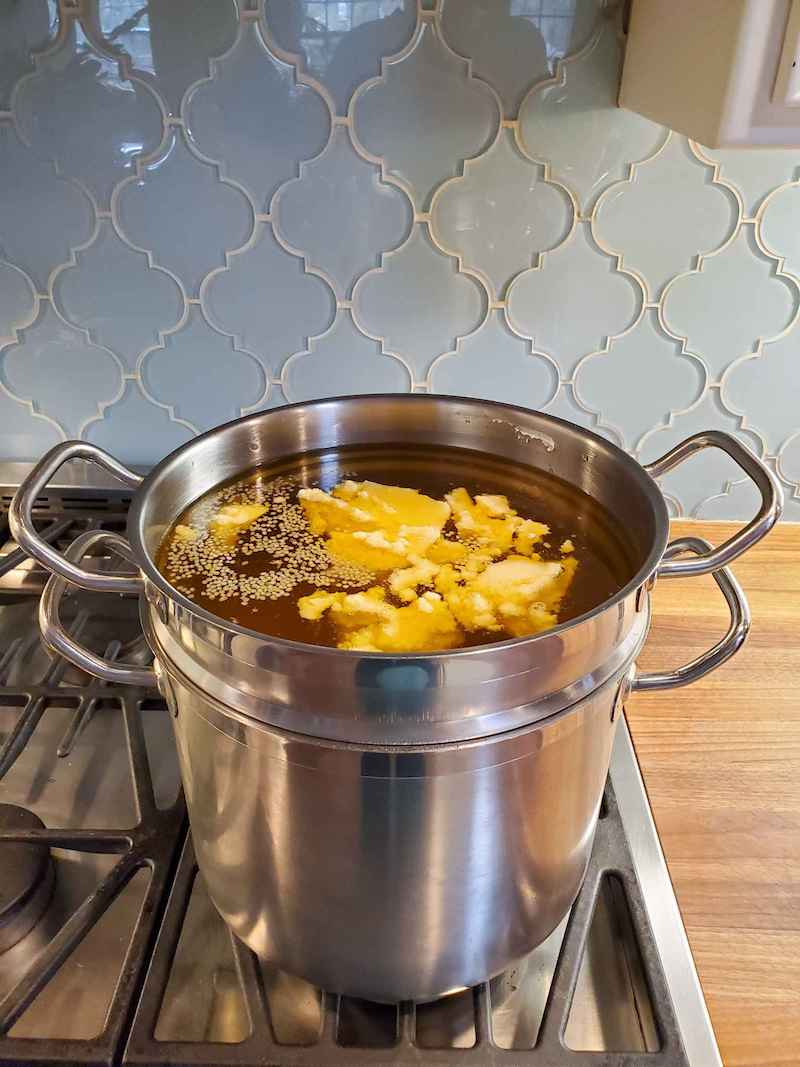 A large double boiler full of oil, beeswax, and shea butter. Some of the shea butter and beeswax pastilles have yet to fully melt and are floating on the top. 