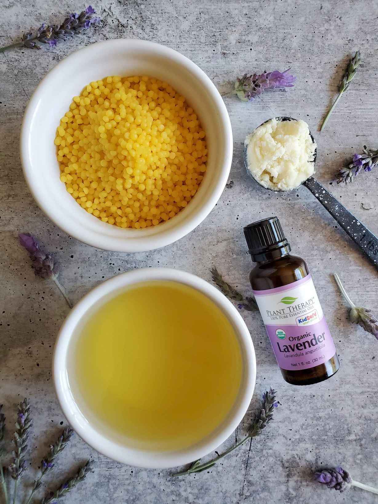 A birds eye view of two white ramekins, one is filled with beeswax pastilles while the other contains oil. A tablespoon measurement is laying off to the side full of shea butter while a bottle of lavender essential oils is laying face up nearby. A number of dried lavender flowers are scattered throughout the scene. 