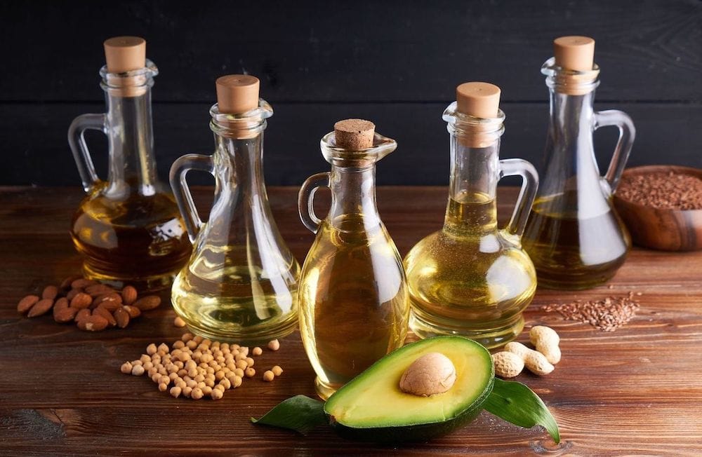 Five glass bottle with cork or rubber tops are arranged in a V-shape. Each one is partially full of oil and next to each bottle contains the item from which the oil was made. Flax, peanut, avocado, almond, along with a less distinguishable nuts or seed. 