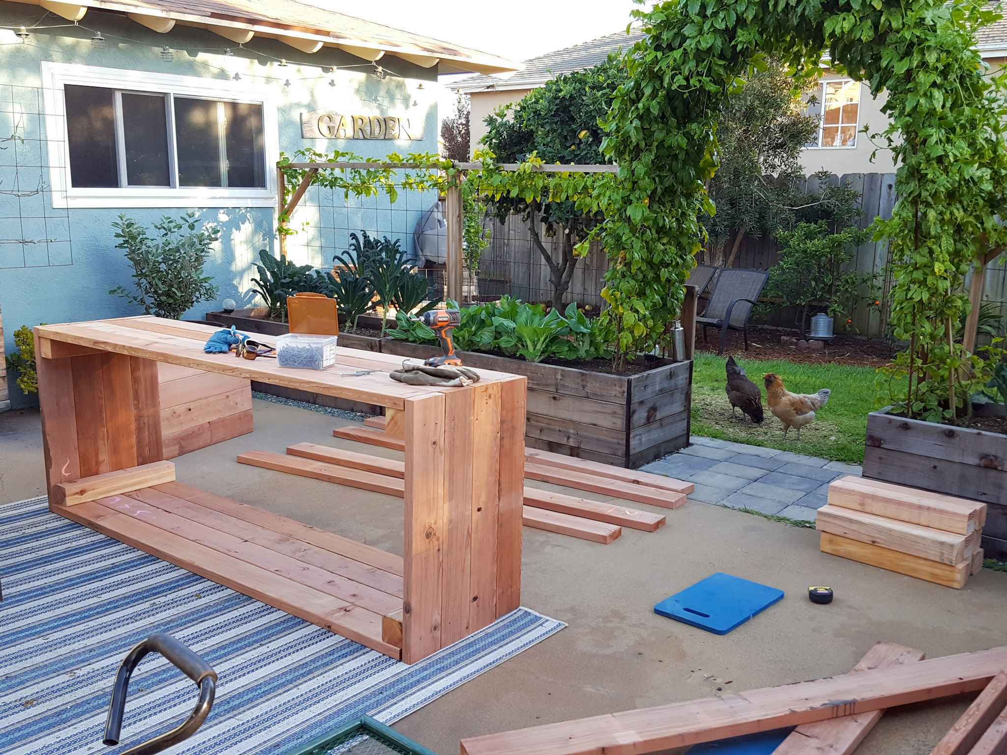 The back patio contains a newly constructed raised garden bed on its side with various pieces and lengths of wood scattered about that are to be used for another bed. There is a drill, gloves, and a box of screws sitting atop the completed raised bed while two chickens look inside into the patio through the arch pathway. 