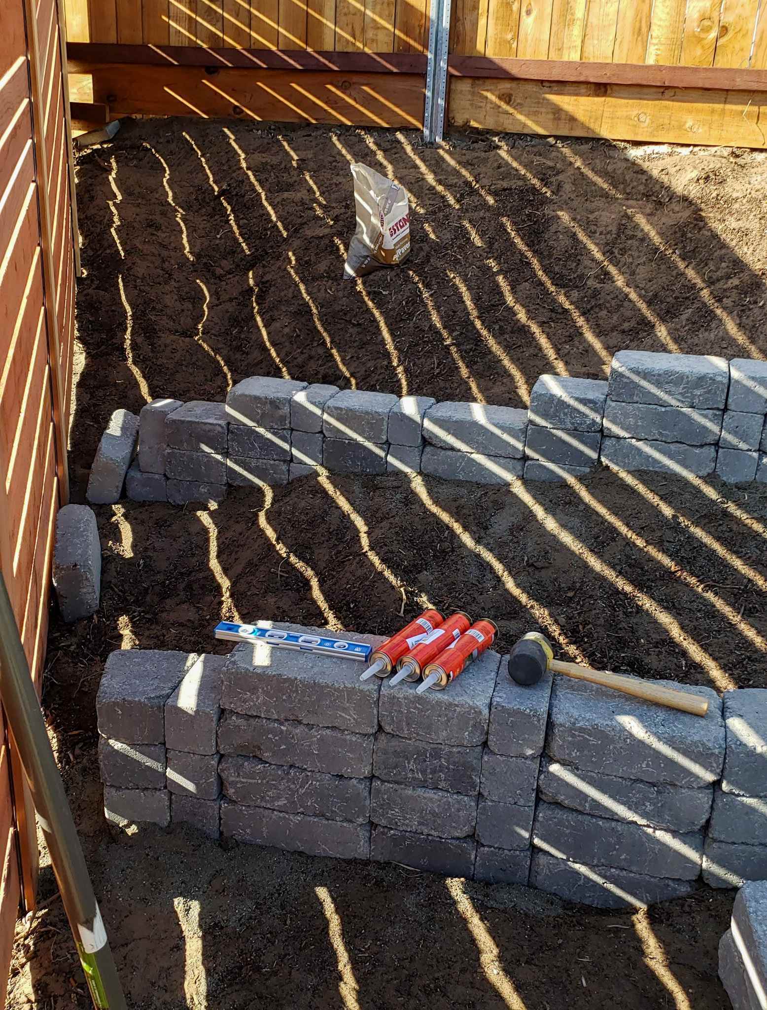 The process of building the stone terrace is underway, dirt is pulled away from each wall of the terrace while the pavers are glued together with strong adhesive.. The tubes of adhesive, a level, and a rubber mallet sit on the lowest formed stone terrace wall. 