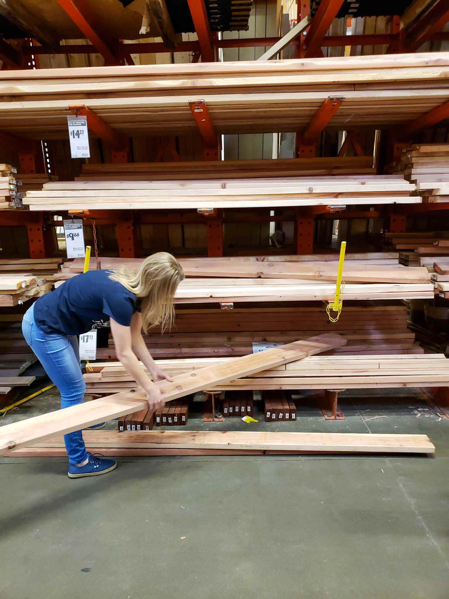 DeannaCat is grabbing a redwood 2x6 board for inspection from a shelf full of boards. There are multiple shelfs with varying sizes and wood materials that spans a height of at least 8 feet. 