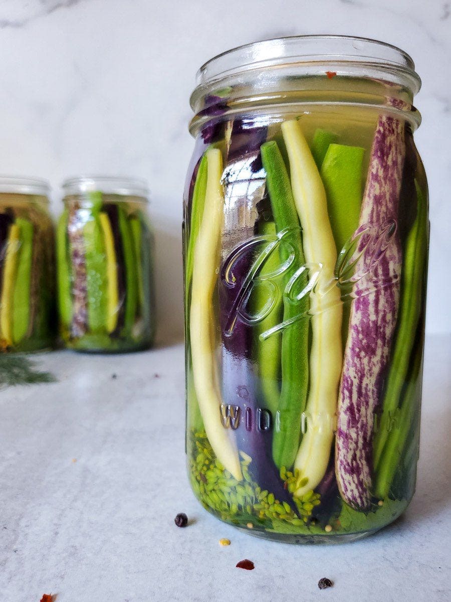 A quart mason jar of pickled green beans is in focus while two other quart mason jars are in the background. Each one is filled with vertically stacked beans of varying colors. 