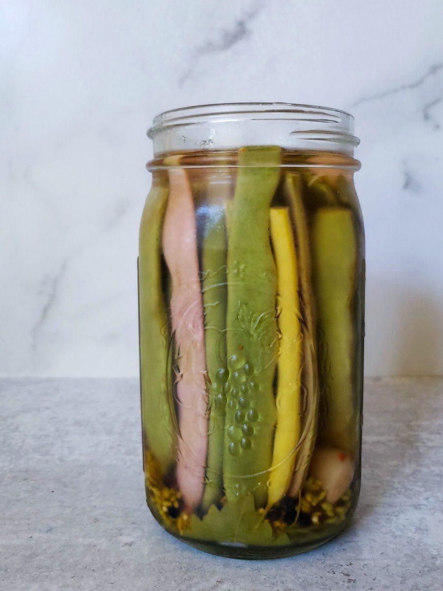 A close up image of quart mason jar filled with neatly stacked veggies that  are sitting inside the jar vertically. The brine is covering the veggies all the way to the tops. Some garlic cloves, a dill head, and grape leaves are visible in the bottom of the jar. 