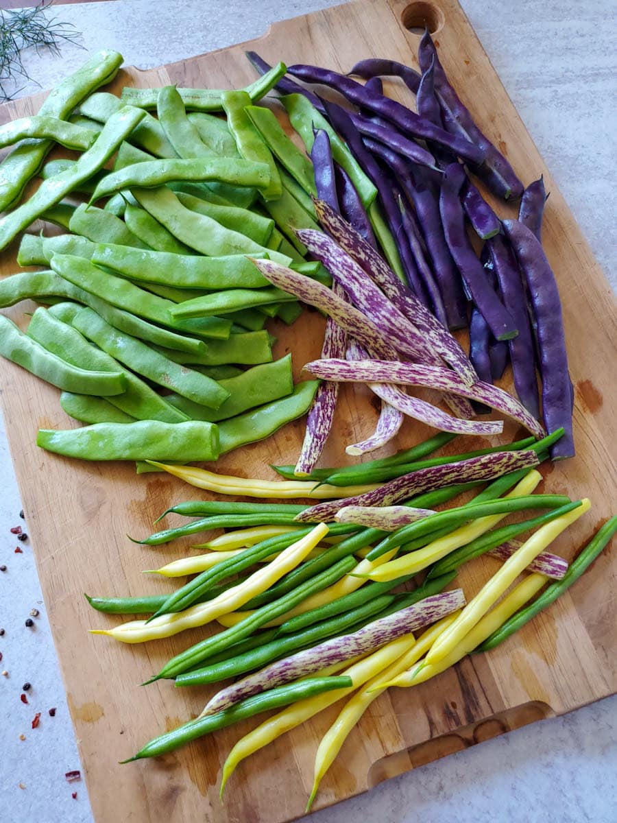 A wood cutting board is covered in prepped veggies. There are a variety of beans on the board, some green, some purples, some yellow, and a few that are white and purple in color. 