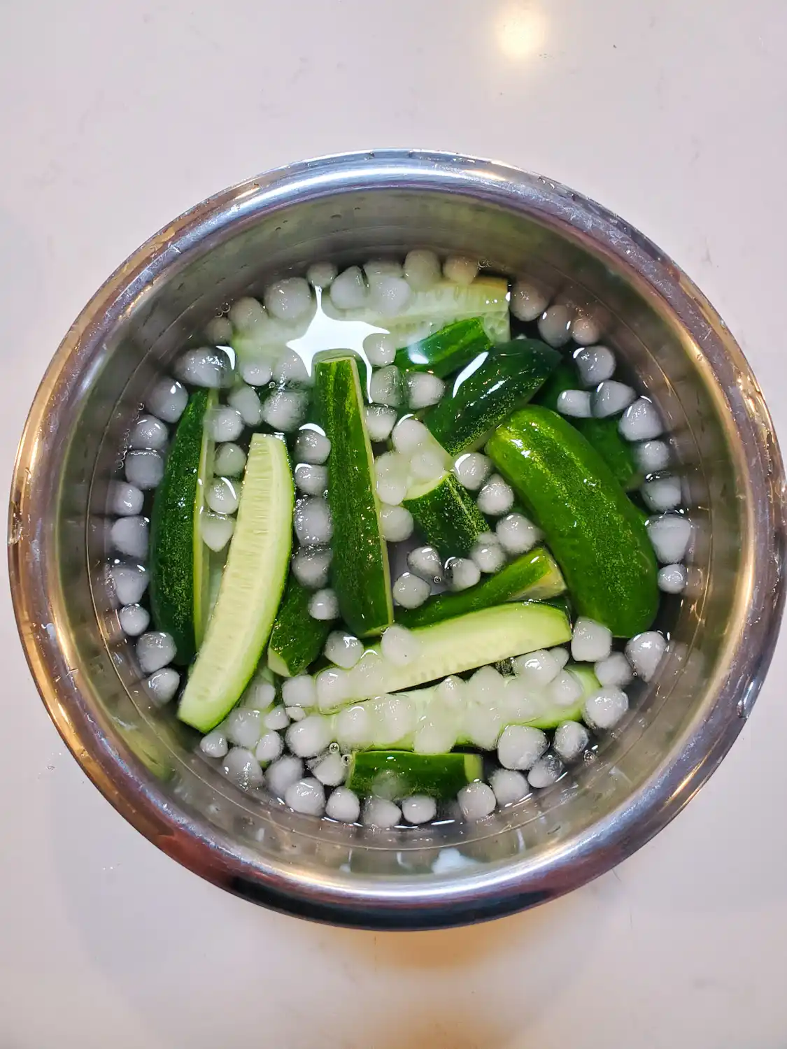 A metal bowl is full of ice cubes floating in water along with halves and quarters of cucumbers floating in the icy water. 