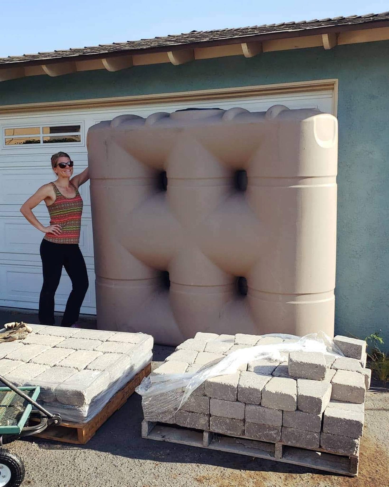 DeannaCat is standing next to the newly purchased 530 gallon slimline rain tank. This is a great tank for rainwater collection in narrow spaces. There are two pallets of pavers and stones sitting in front of them, though these are for a separate project unrelated to the rain water tank. 