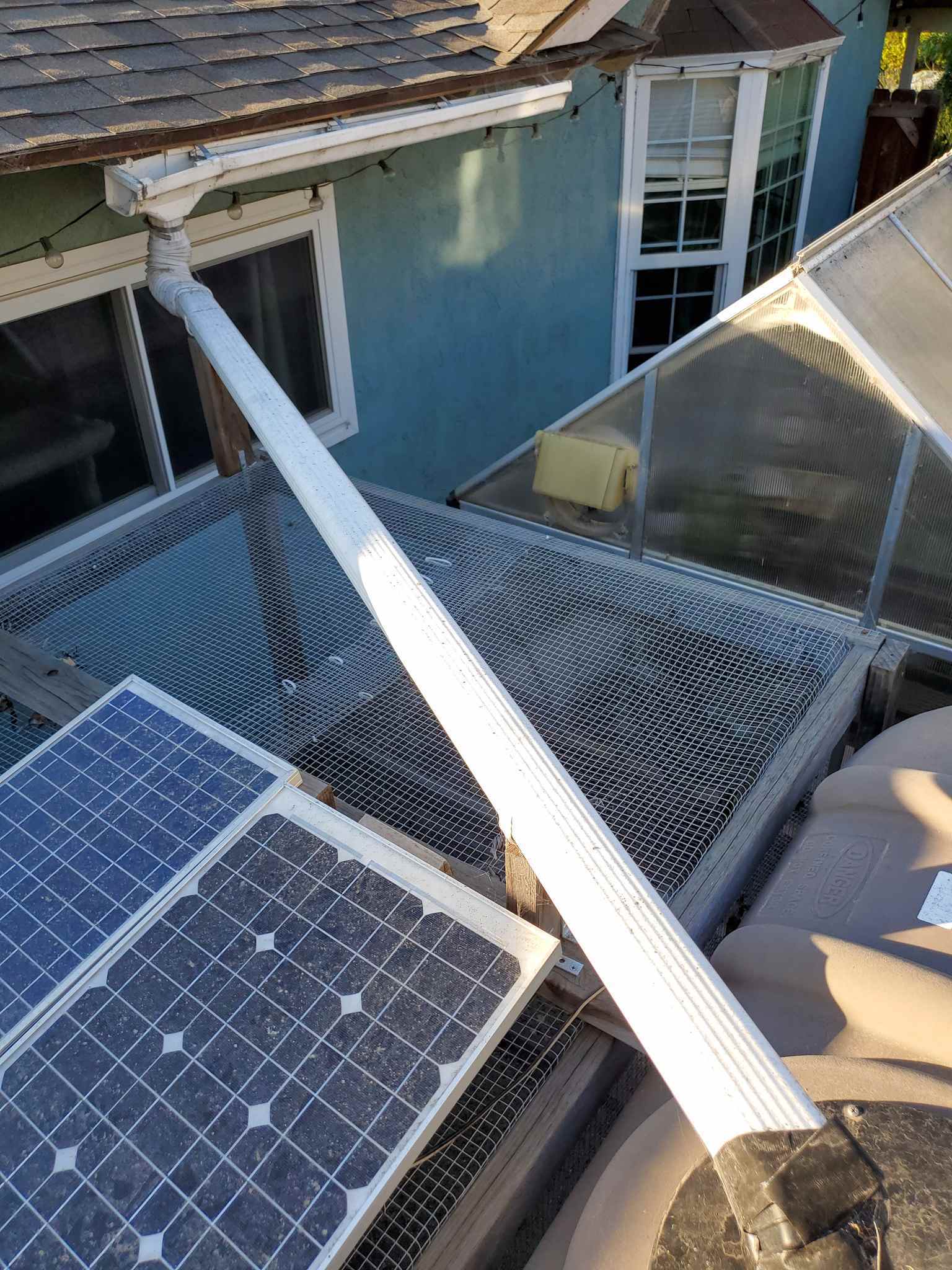 A view from the top of a rain tank, it shows a downspout connected to a gutter, traversing a chicken run and connected to the top of the rain tank. There are solar panels on top of the run next to the tank which power fans that are inside the greenhouse which is visible from the tank.