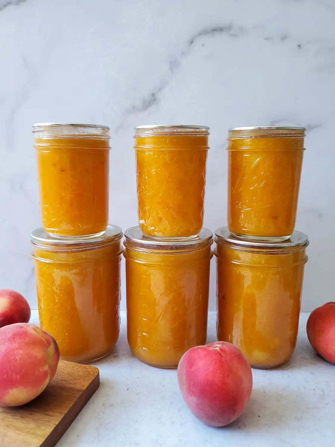 Three half pint jars are lined up in a straight line, each one sitting on the top of a pint jar, all of the jars contain low sugar peach jam. A number of whole peaches are scattered around the jam jars. 