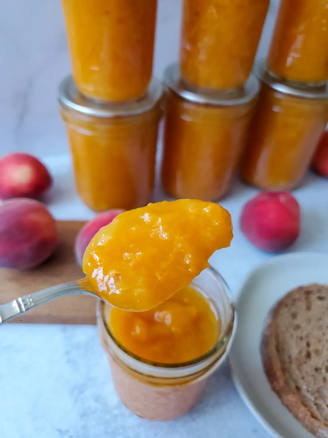 A half pint mason jar is full of low sugar peach jam, a spoon is suspended above the jar after scooping out a spoonful of the jam. Beyond lies a few jars of the jam along with a number of whole peaches scattered around the area. 