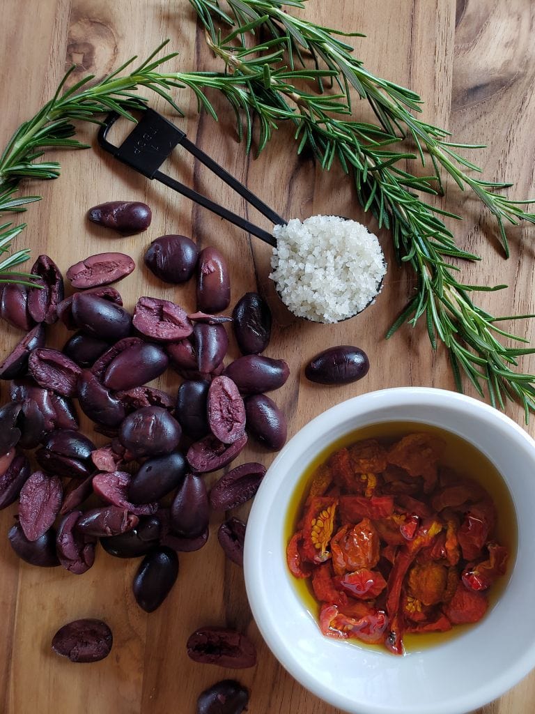 A birds eye view of a wooden cutting board with rosemary sprigs, a heaping tablespoon of celtic grey salt, kalamata olives sliced in halves, and a white ramekin of of sun-dried tomatoes submerged in olive oil.