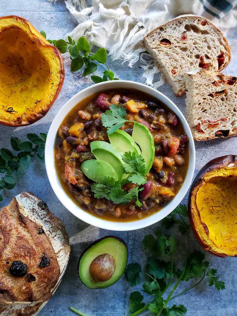 The roasted pumpkin chili is shown garnished with avocado slices and cilantro leaves. Surrounding the bowl in a decorative fashion are halves of roasted pumpkin, slices of sourdough bread as well as half of the remaining loaf, half an avocado, sprigs of cilantro, and sprigs of oregano. 