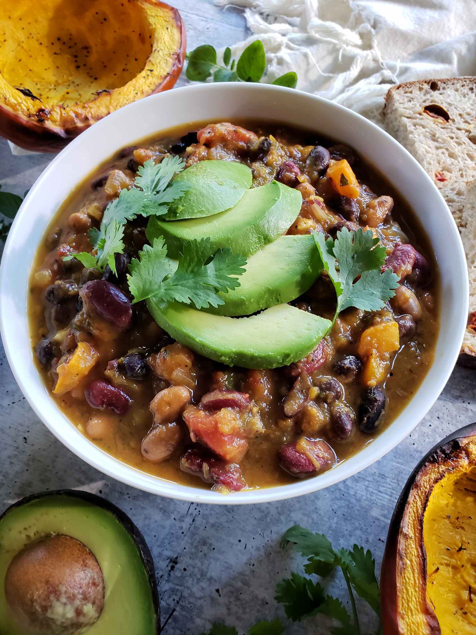 The finished roasted pumpkin chili is shown inside a white ceramic bowl. The beans, tomato, and pumpkin are visible amongst the dark brownish red of the chili base. The chili has been garnished with avocado slices and a few leaves of cilantro. There is half of a roasted pumpkin above the bowl, while a piece of sourdough bread is slightly visible to the right. Another roasted pumpkin half is partially visible to the lower right of the bowl and half an avocado with the pit remaining is visible to the lower left. 