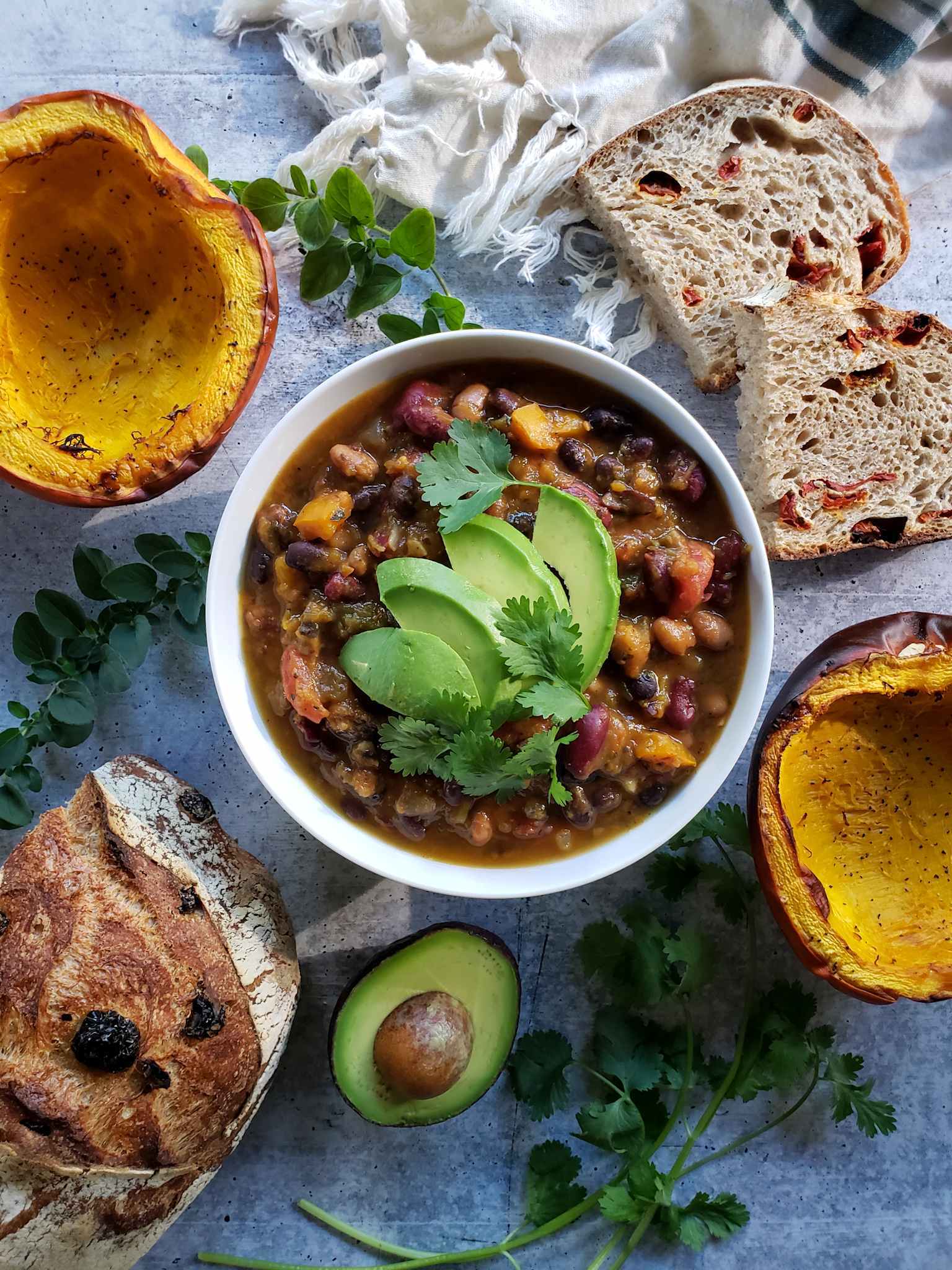 The roasted pumpkin chili is shown garnished with avocado slices and cilantro leaves. Surrounding the bowl in a decorative fashion are halves of roasted pumpkin, slices of sourdough bread as well as half of the remaining loaf, half an avocado, sprigs of cilantro, and sprigs of oregano. 