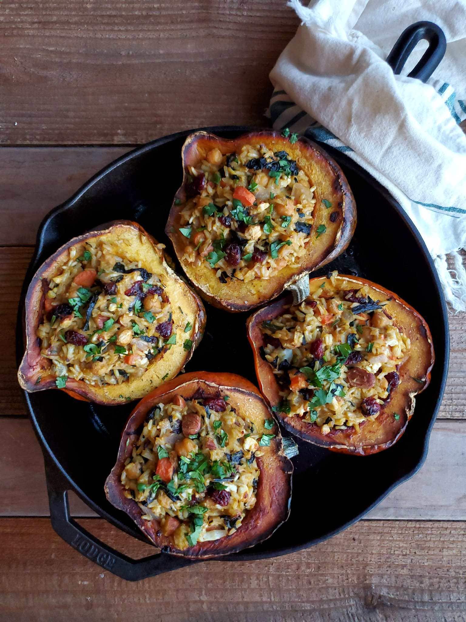 Four halves of biryani stuffed squash sitting in a cast iron pan after it has be baked in the oven. Each squash half is garnished with cilantro, while the dried fruit, nuts, and garbanzo beans are visible amongst the rice stuffing. 