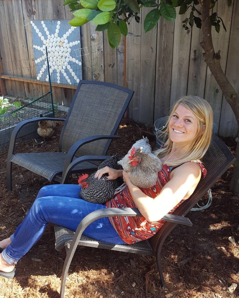 Deana is sitting on a chair in the garden, smiling up at the camera as she holds two chickens. One grey one is cradled up on her chest, and a black and white one is nestled in her lap.  