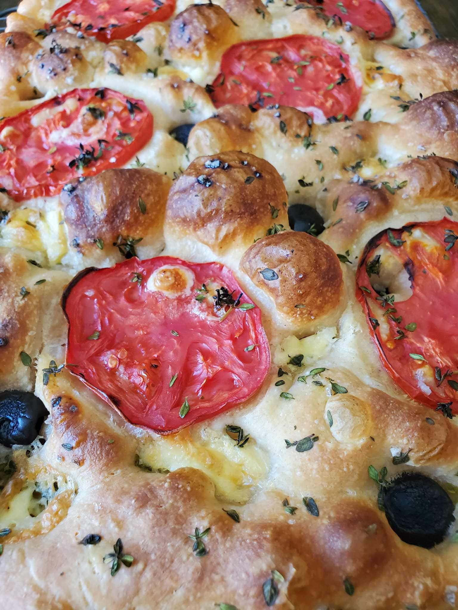 A close up of baked sourdough focaccia, with a light brown bubbly surface, flecks of little green thyme leaves, slices of round tomatoes, and black olives pressed into the dimples on top of the dough.