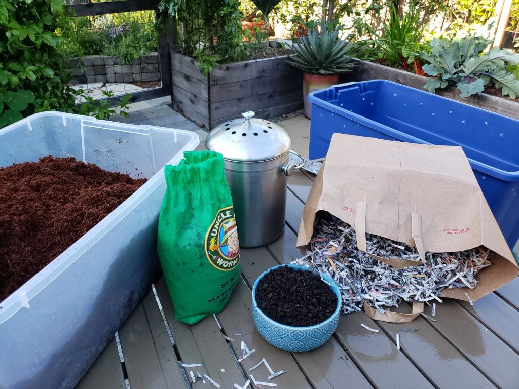 All of the supplies needed to make worm compost. A tub of hydrated coco coir, a bag of compost worms, a plastic bin with air holes, shredded paper, and fruit or vegetable material as additional food for the worms. 